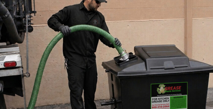 A worker in dark clothing and gloves uses a large green hose to pump grease from a black outdoor container labeled “Grease Trap Services” against a tan wall.